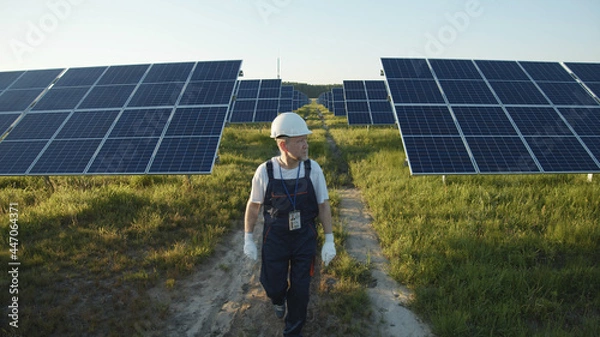 Obraz Adult engineer in uniform walking across solar battery panels on green sunny field. Futuristic technology innovation. Ecology and environment concept. Sunlight power. Eco-friendly system.