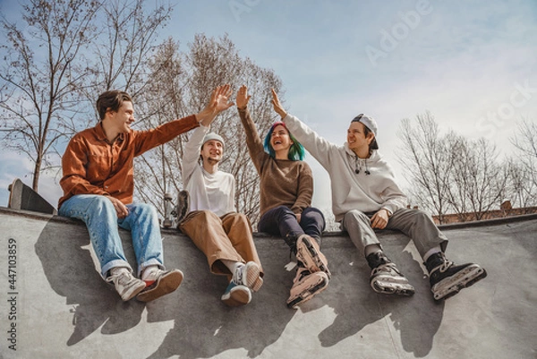 Fototapeta Four rollerbladers and skateboarders, three boys and one girl, sit on a ramp in a city skatepark and give five to each other, cheering up after a fun workout