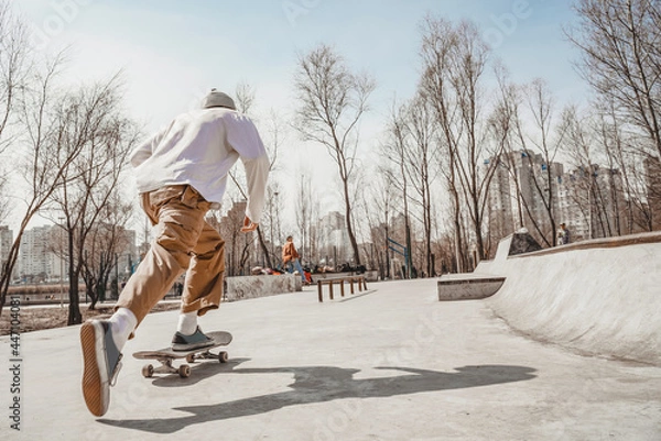 Fototapeta Skater guy rides a skateboard very quickly, pushing off the ground with his right foot, goes to his skater friends. The sun reflects the shadow of a skater on the gray surface of a skatepark.