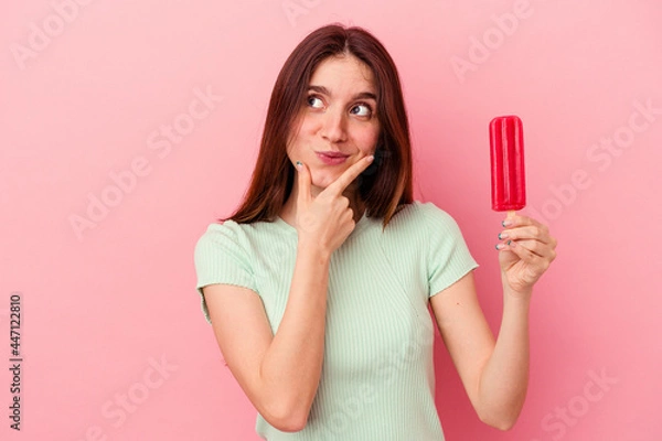 Obraz Young caucasian woman holding an ice cream isolated on blue background looking sideways with doubtful and skeptical expression.