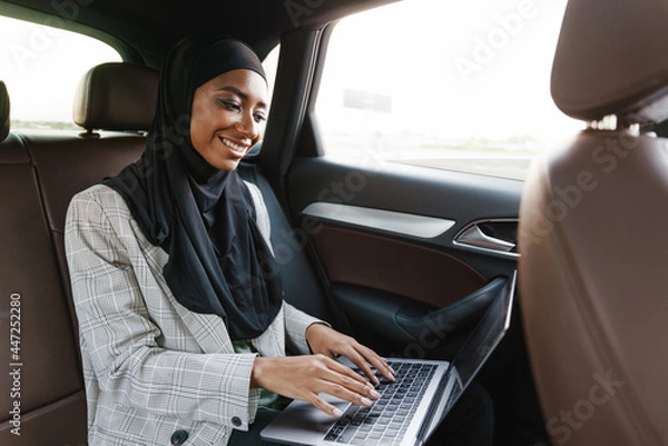 Fototapeta Black muslim woman working with laptop while going in car