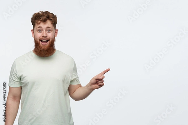 Fototapeta indoor shot of young bearded redhead guy, wears blank t-shirt, pointing with a finger at right side copy space, smiling and has amazed facial expression. isolated over white background.