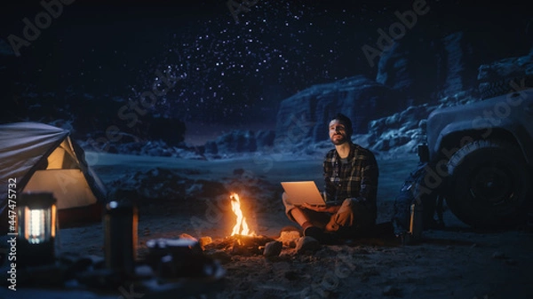 Fototapeta Night Tent Camping in Canyon: Male Traveler Uses Laptop Computer Sitting by Campfire. Man on Digital Remote Work, e-shopping, ecommerce, Using Internet, Social Media Posting on Vacation Trip 