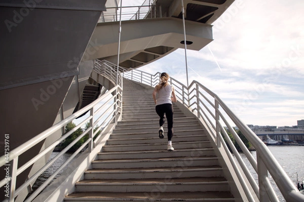 Obraz A young woman runs up the stairs of the bridge. Power training. Running in the evening in the city