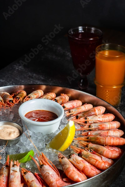 Fototapeta Royal shrimp on ice. A plate with cold fresh prawns, ice, lemon, lime and sauce. Sea buckthorne morse on the table. Low key. Vertical orientation.