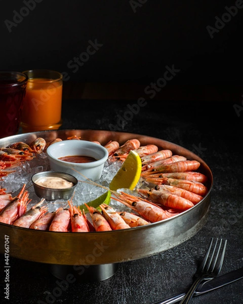 Fototapeta Royal shrimp on ice. A plate with cold fresh prawns, ice, lemon, lime and sauce. Sea buckthorne morse on the table. Low key. Vertical orientation.