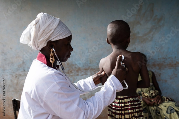 Obraz Young smiling doctor in a clean white uniform auscultating a little boy's lungs sounds with a modern stethoscope on his bare back