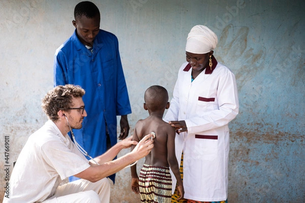 Obraz White medical volunteer auscultating the lungs of a skinny little African boy under the supervision of the responsible pediatrician and a nurse both dressed in their hospital uniforms