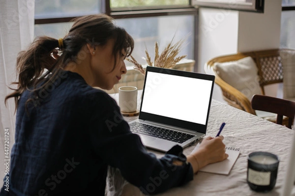 Fototapeta View from back of woman using laptop and writing in notebook. Mockup White screen laptop, computer