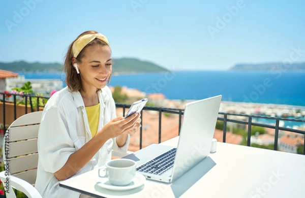 Fototapeta Young elegant woman wearing wireless headphone looking at smartphone with smile while sitting at balcony with seaview on resort. girl watching video, chatting in social network