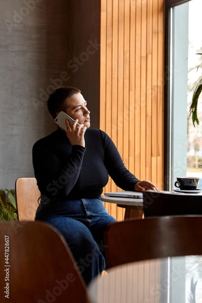 Fototapeta Young girl with short hair and nose piercing sits in a cafe. A woman is talking on the phone and work at a tablet. The concept of freelancing and remote work.