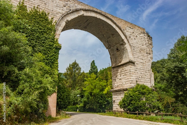Fototapeta The ruins of the Roman arch bridge of Augustus, in Narni, Terni, Umbria. The remains of the bridge over the Nera river. The big and ancient stone arch, against the blue sky. Trees and dense vegetation