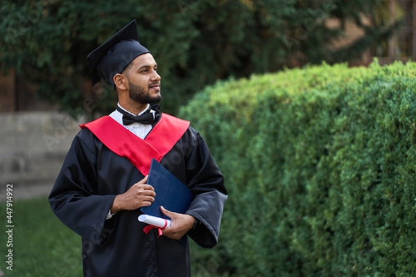 Fototapeta Portrait of handsome indian graduate student in graduation glow with diploma looking forward for copy space text
