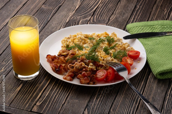 Fototapeta Orange juice and scrambled eggs with bacon and tomatoes on a wooden table