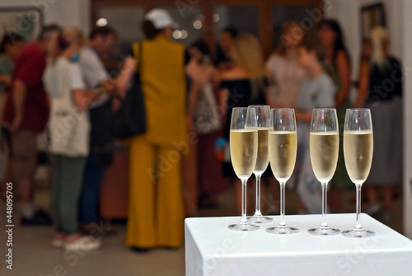 Fototapeta Glasses with wine on the background of the exhibition of paintings. People blurred in the background. Art salon with white walls.