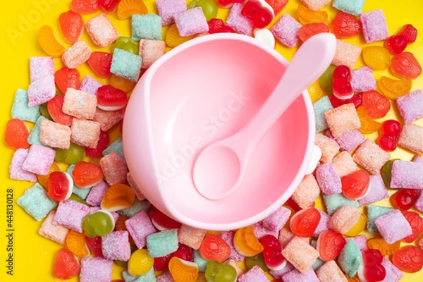 Fototapeta Children's tableware silicone bowl with spoon. Serving baby food, kid dishware.Pink silicone bowl and spoon are in a heap of sweets and marmalade. bright, colorful photo flatly