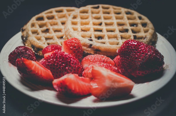 Fototapeta The chocolate chips warm waffle with fresh strawberries on the side for breakfast.. White plate. Black background. Selective focus.
