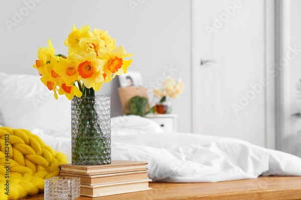 Fototapeta Vase with narcissus flowers and books on bedside bench in room