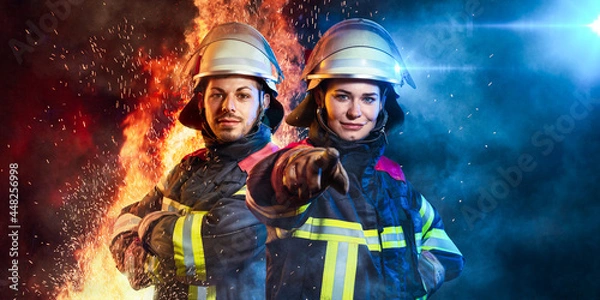 Fototapeta Ein Feuerwehrmann und eine Feuerwehrfrau stehen zusammen vor Feuer und Blaulicht
