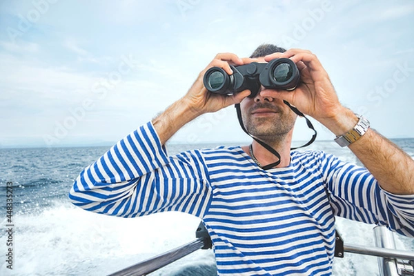 Fototapeta young man, sailor in striped jacket - vest with stubble sits on boat in sea and looks ahead through binoculars. sea navigation, summer tourism. travel content, selective focus