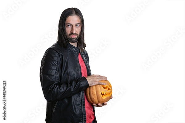 Fototapeta A man with a beard and long hair in a black jacket with a pumpkin in his hands posing on a white background. Various poses and emotions. Halloween concept. 