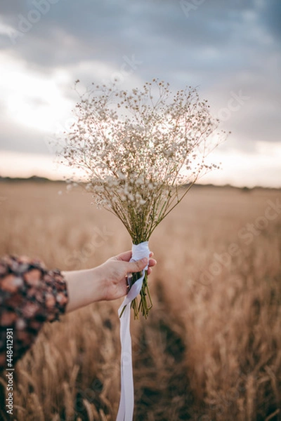 Obraz a girl in a sundress, in a field holding a hat and flowers in her hands
