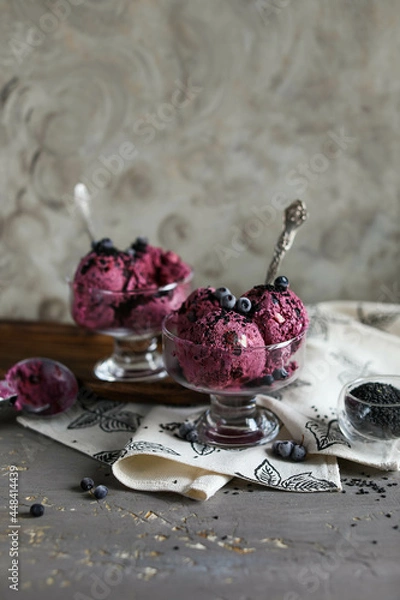 Fototapeta Blueberry ice cream, garnished with berries and black sesame seeds, in glass bowls with spoons on a dark gray table. Cold summer desserts.