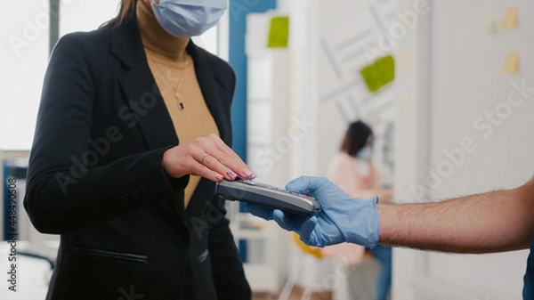Fototapeta Closeup of businesswoman paying takeaway food order with credit card using POS contactless service during takeout lunchtime. Delivery man with medical face mask and gloves giving fresh food meal
