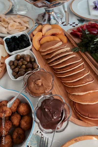 Fototapeta Large selection of breakfast food on a table with pancakes, fruits, bakery, cold meats, cheese and jam, olives and walnuts served. Viewed from above.