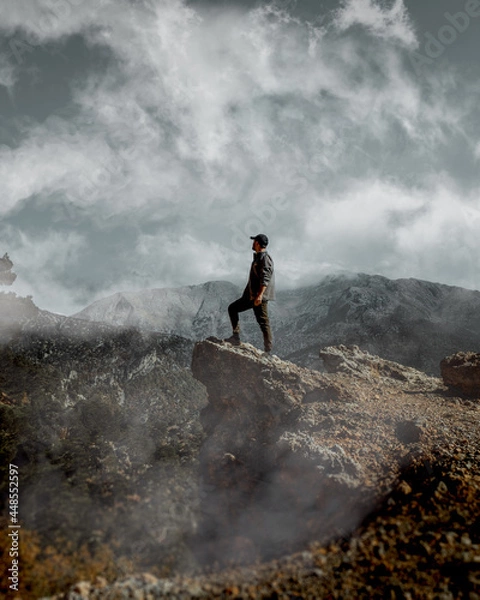 Fototapeta A person standing on the edge of a cliff with sky, clouds and mountains in the background.