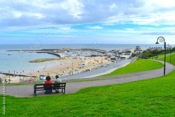 Obraz Couple sat on bench overlooking the Cobb in Lyme Regis surrounded by beach and greenery Many activities are available starting with kids trampoline park right on the sand mini golf and paddle boarding