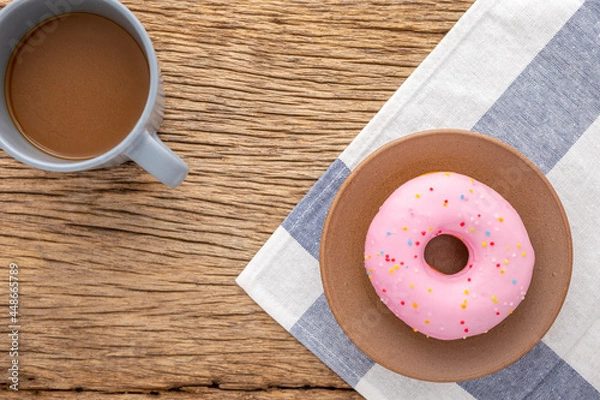 Fototapeta pink strawberry donut in brown terracotta plate on white and blue napkin beside coffee on rustic natural wood texture background, easy meal for break time, top view