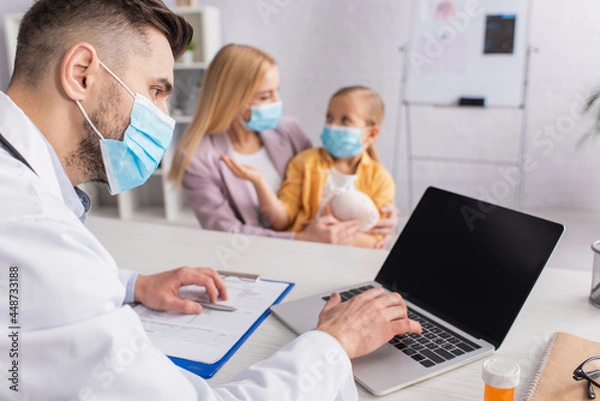 Fototapeta Pediatrician in medical mask using laptop near pills and family in clinic