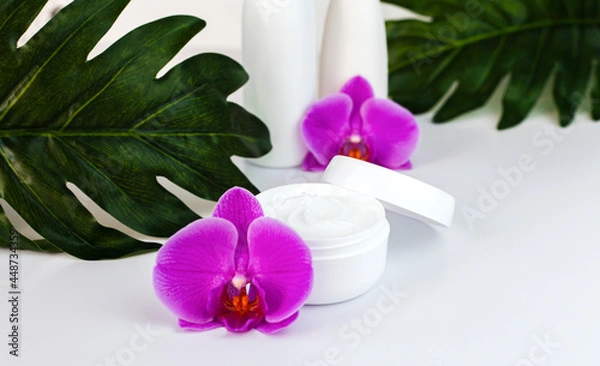 Fototapeta Moisturizing cream in a white jar with orchid flowers on white background. Natural cosmetics concept. Face and hand care cosmetics. Close-up. Selective focus.