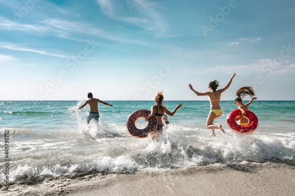 Obraz Happy cheerful friends resting on a beautiful beach at sunny day and running into the sea with inflatable rings. Tropical vacations concept