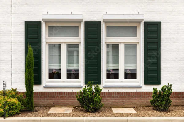 Fototapeta Symmetrical facade of a white modern house with an orange roof in Thorn, The Netherlands, known for its white houses and buildings. Green shutters next to the windows, greenery and plants