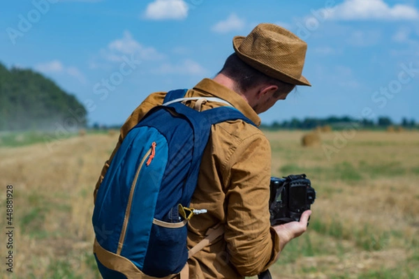 Fototapeta Traveler photographer with a camera in his hand against the background of a field and haystacks.