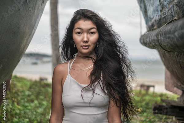 Fototapeta Portrait of beautiful young mongol woman in white dress standing between two old wooden ships. Brunette long curly hair. Looking at camera with copy space. Romantic portrait.