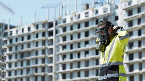 Obraz An employee of the gas and environmental inspection puts on a gas mask against the background of an industrial unfinished building