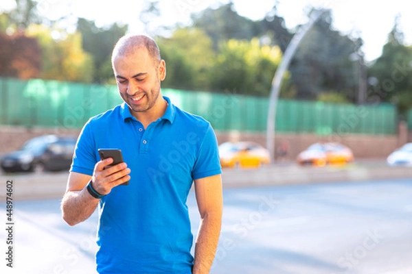 Fototapeta Cheerful and confident young man checking his e-mails and looking to his smart phone in an urban. Includes copy and text space.