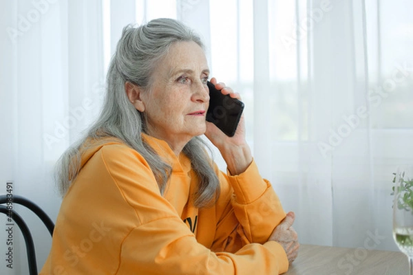 Fototapeta Beautiful old grandmother with grey hair and face with wrinkles is using smartphone, talking with someone and sitting at the table at home on window background, mother's day, happy retirement