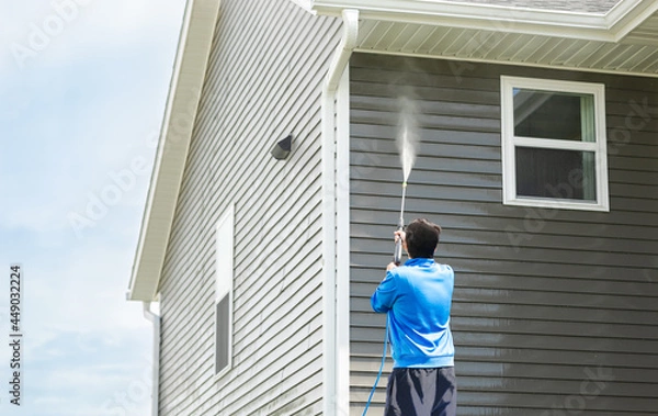 Fototapeta Man in blue jacket clean dusk and dirt from exterior siding and under roof by high-pressure nozzle spray with water soap cleaner, weekend chore on sunny day. Cleansing and maintenance service concept.