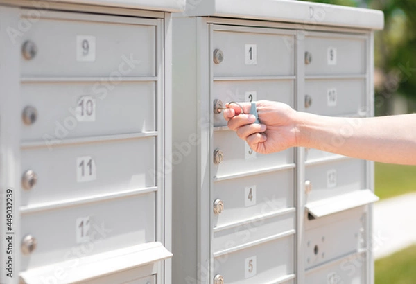 Fototapeta Person’s hand holding key unlock-lock mailbox by house number, selective focus. Receiver get mail and small parcel from row of outdoor metal storage, depth of field. Man close residential letterbox.