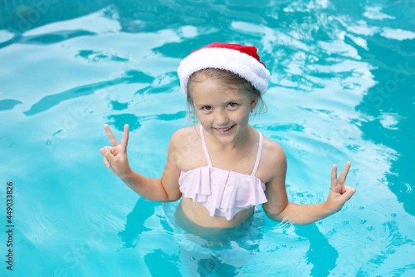 Fototapeta a beautiful happy girl in a Santa Claus hat in a pink swimsuit is standing in the pool for the new year