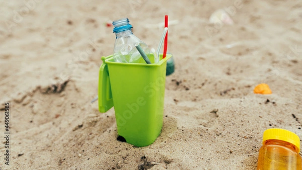 Fototapeta Small toy waste container on the sand beach with plastic garbage
