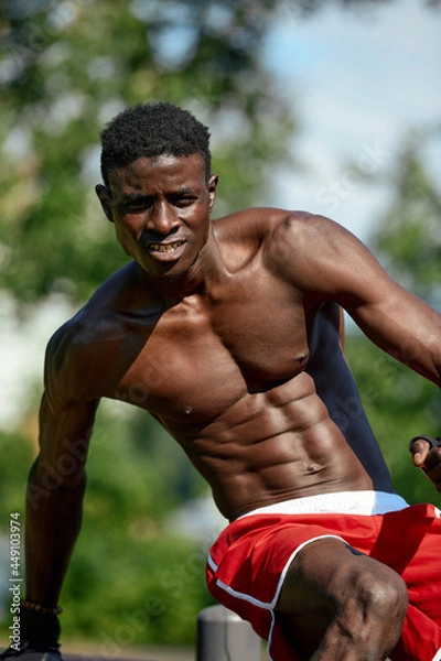 Fototapeta Muscular young man exercising at the sports field. African man looking to the side while doing horizontal bar exercises. Shirtless male model exercising outdoors.