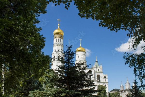 Fototapeta The high Kremlin white bell tower with a golden dome Ivan the Great in a natural frame made of tree branches with green leaves on a sunny summer day and blue sky in Moscow Russia