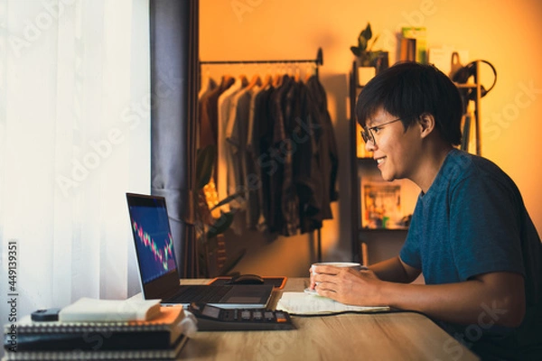 Fototapeta Asian young man 30s looking stock tickers or graphs cryptocurrency trading platforms on the laptop holding coffee ready start to hard work. work from home, financial at the home office in the morning