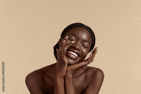 Fototapeta Portrait close up of beautiful african girl with closed eyes. Smiling young woman touch her clean face. Concept of face skin care. Isolated on beige background. Studio shoot
