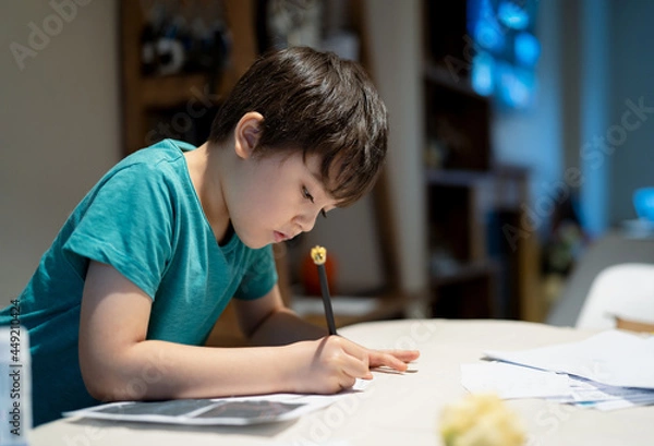 Fototapeta Portrait of school kid siting on table doing homework, Child boy holding pencil writing on paper, Homeschooling , back to school concept